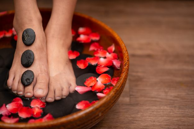 A pair of feet sit in a pail of water with flower petals and stones